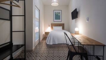 Habitación de hotel estrecha con cama, mesita de noche, silla y lámpara colgante.