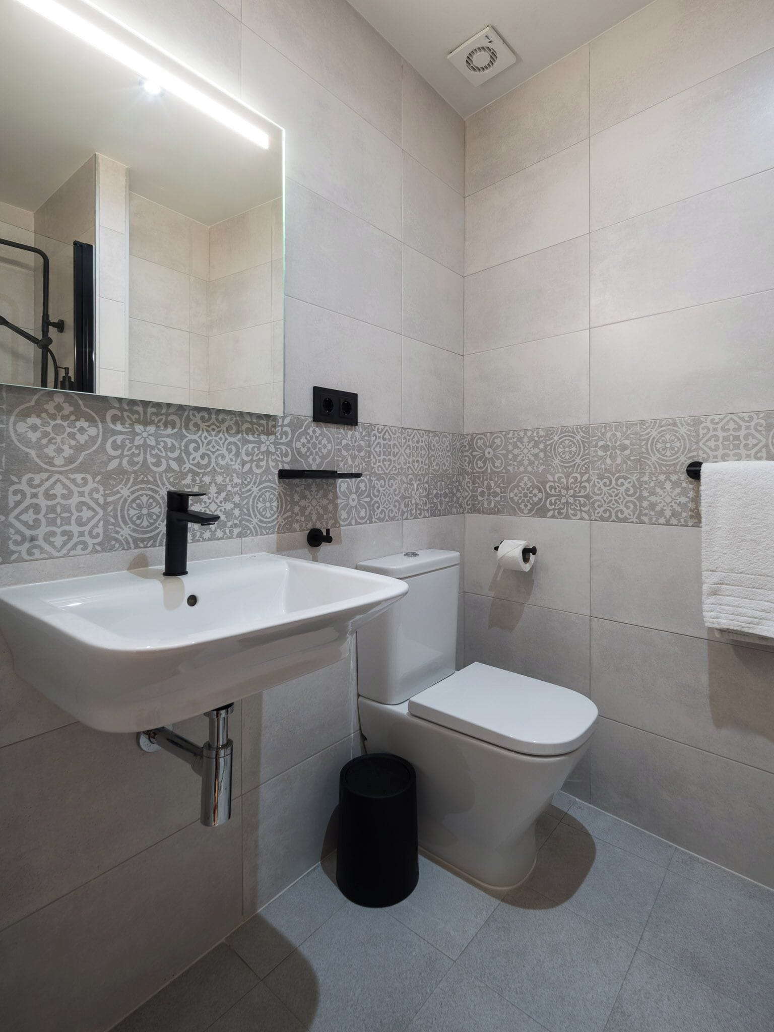 Un baño con lavamanos blanco, grifo negro, inodoro blanco y azulejos estampados.