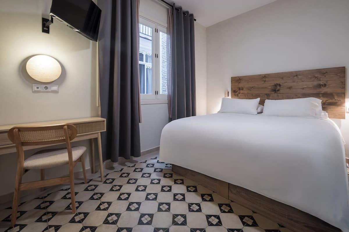 Een hotelkamer met een bed, een bureau met stoel, een aan de muur bevestigde televisie en een raam met gordijnen.