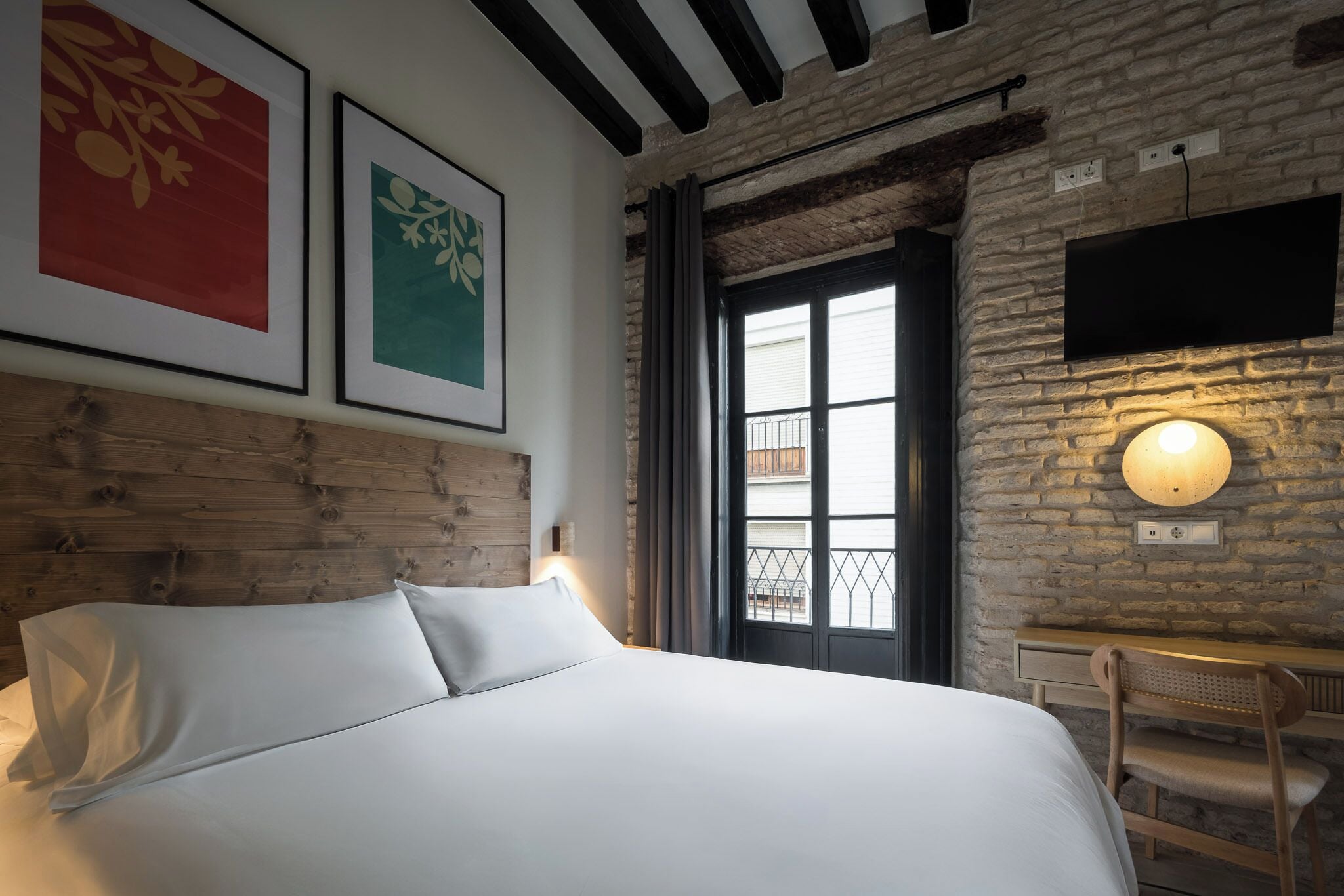 A bed with white linens, a wooden headboard, a desk with a chair, a wall-mounted TV, and framed artwork.
