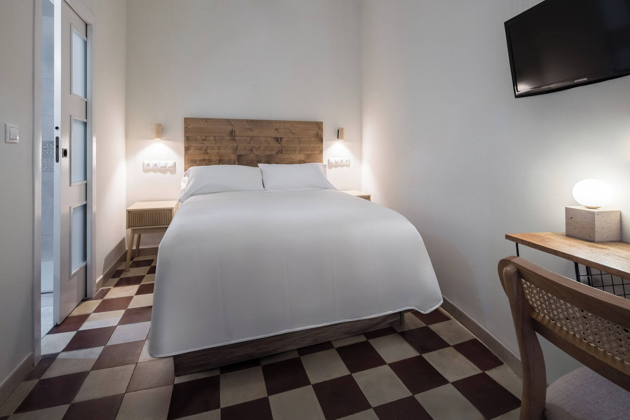 Quarto de hotel com piso xadrez, cabeceira de madeira, cama com roupa de cama branca, abajures, televisão de tela plana e uma pequena mesa com luminária.