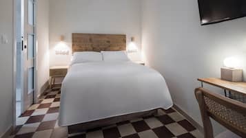 Quarto de hotel com piso xadrez, cabeceira de madeira, cama com roupa de cama branca, abajures, televisão de tela plana e uma pequena mesa com luminária.