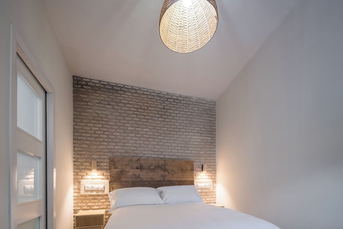 Een slaapkamer met een groot bed, een muur met baksteenpatroon, een houten hoofdeinde, twee bedlampen en een kamerkleed met dessin.