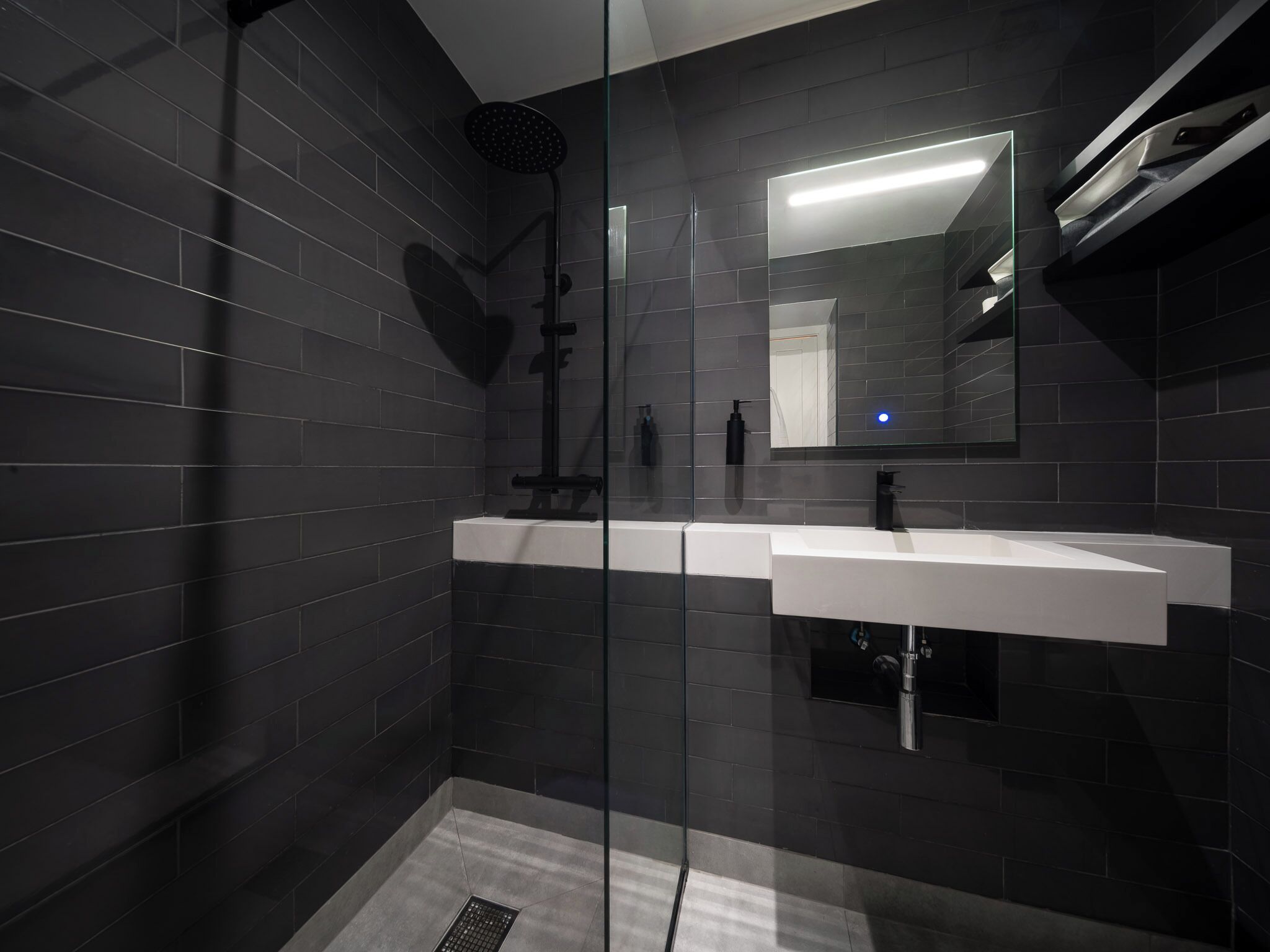 A modern bathroom with dark tiles, a glass shower enclosure, and a large mirror.