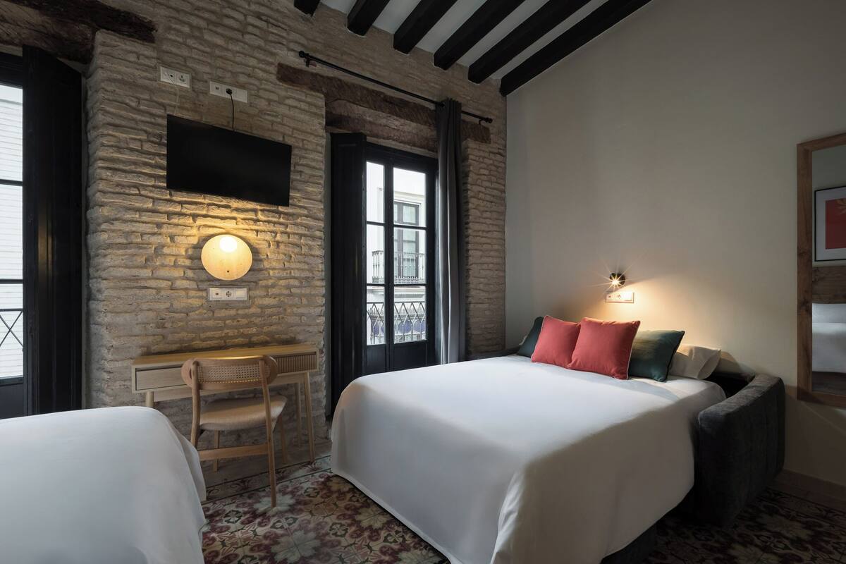 Een kamer met twee bedden, een bureau, een stoel, een aan een bakstenen muur bevestigde televisie en een balkon met een hekwerk.