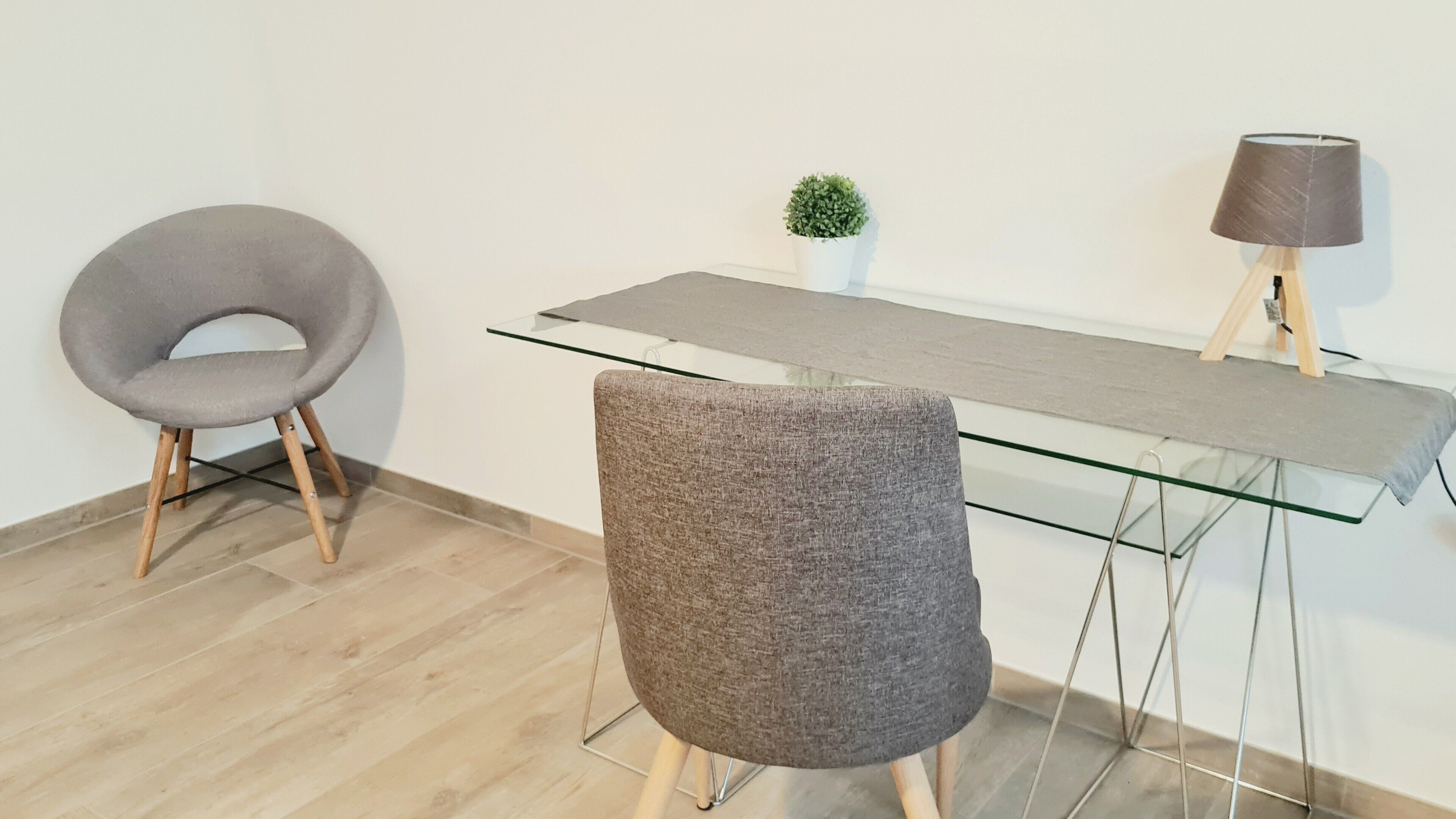 Un espace de bureau moderne avec un bureau en verre, deux chaises grises, une petite plante en pot et une lampe posée sur un support en bois.