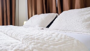 Ein Bett mit weißer, strukturierter Decke und Kissen, braunen Vorhängen und einem Nachttisch mit Lampe.