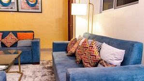 Ein Wohnzimmer mit einem blauen Sofa, gemusterten Kissen, einem Glastisch und einer Lampe.