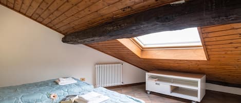 Una habitación con techo inclinado, un tragaluz, una cama con colcha azul, un radiador blanco y un mueble blanco para guardar objetos.