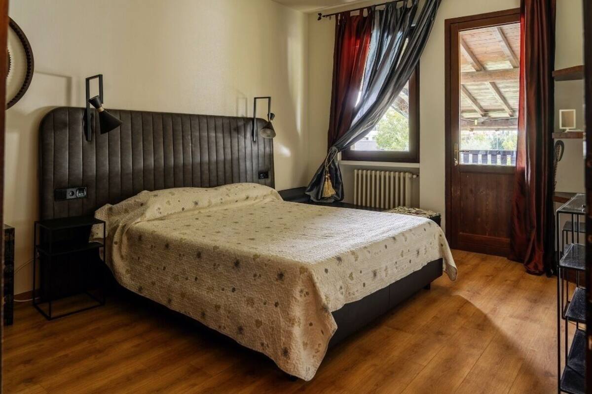 A bedroom with a bed, bedside table, lamp, window with curtains, radiator, and wooden floor.
