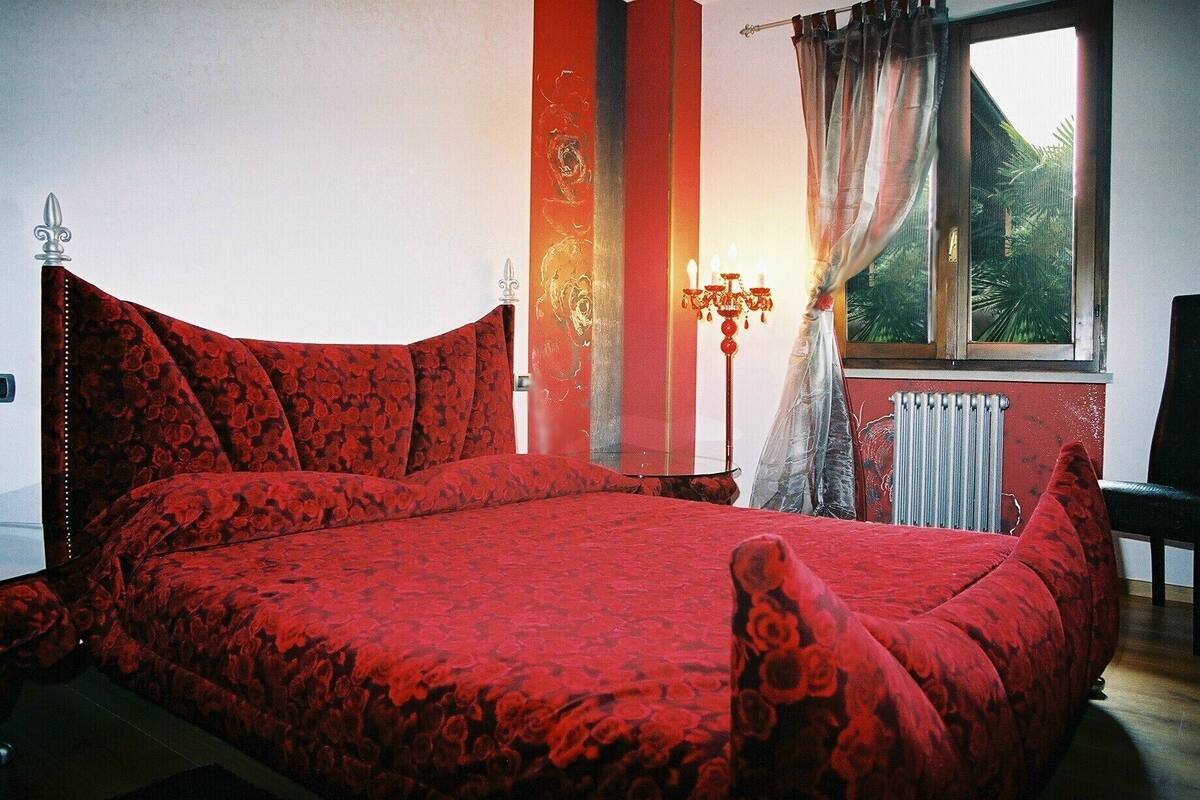 A bedroom with a large bed, a radiator, a window with curtains, and a nightstand with a lamp.