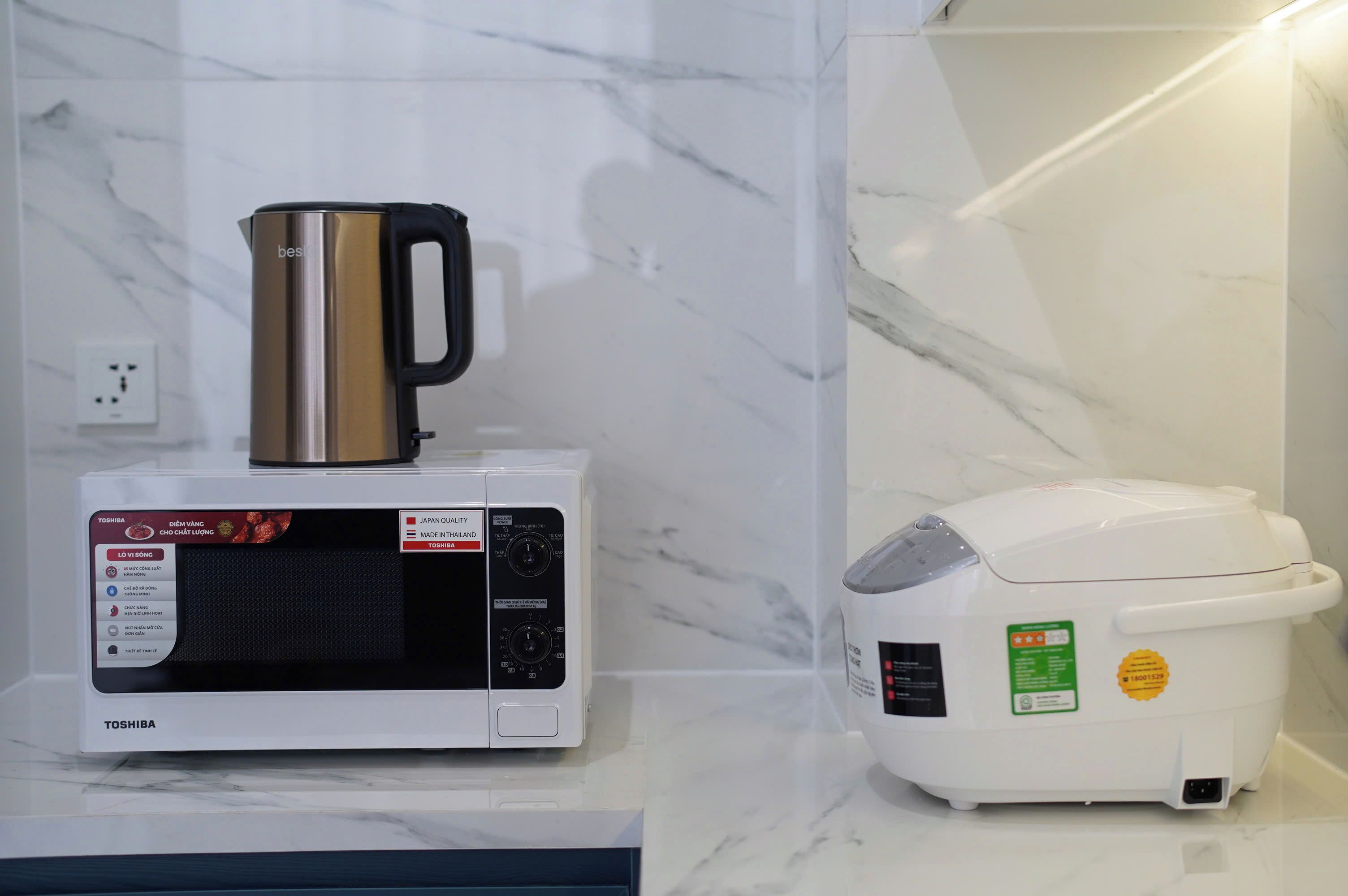 A microwave with a coffee pot on top, a rice cooker, and a wall socket.