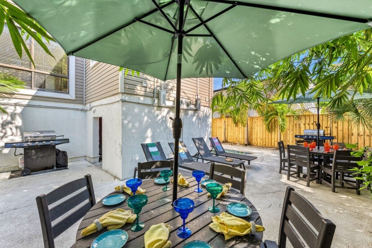 Un patio al aire libre con una mesa redonda dispuesta para comer, rodeada de sillas y una sombrilla que proporciona sombra.