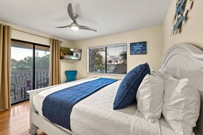 Un dormitorio con cama, ventilador de techo, televisión, ventana con persianas y balcón.