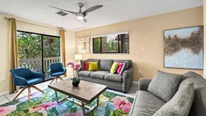 A living room with a sofa, two armchairs, a coffee table, a ceiling fan, and a large window with a view of greenery.