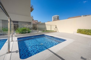 A swimming pool with clear blue water, surrounded by a white building with a glass enclosure and a view of a tall building in the background. - Free Housekeeping Visits During Your Stay - Stayshort - Comfortable Studio in JVC That Sleeps 2 (Dubai)
