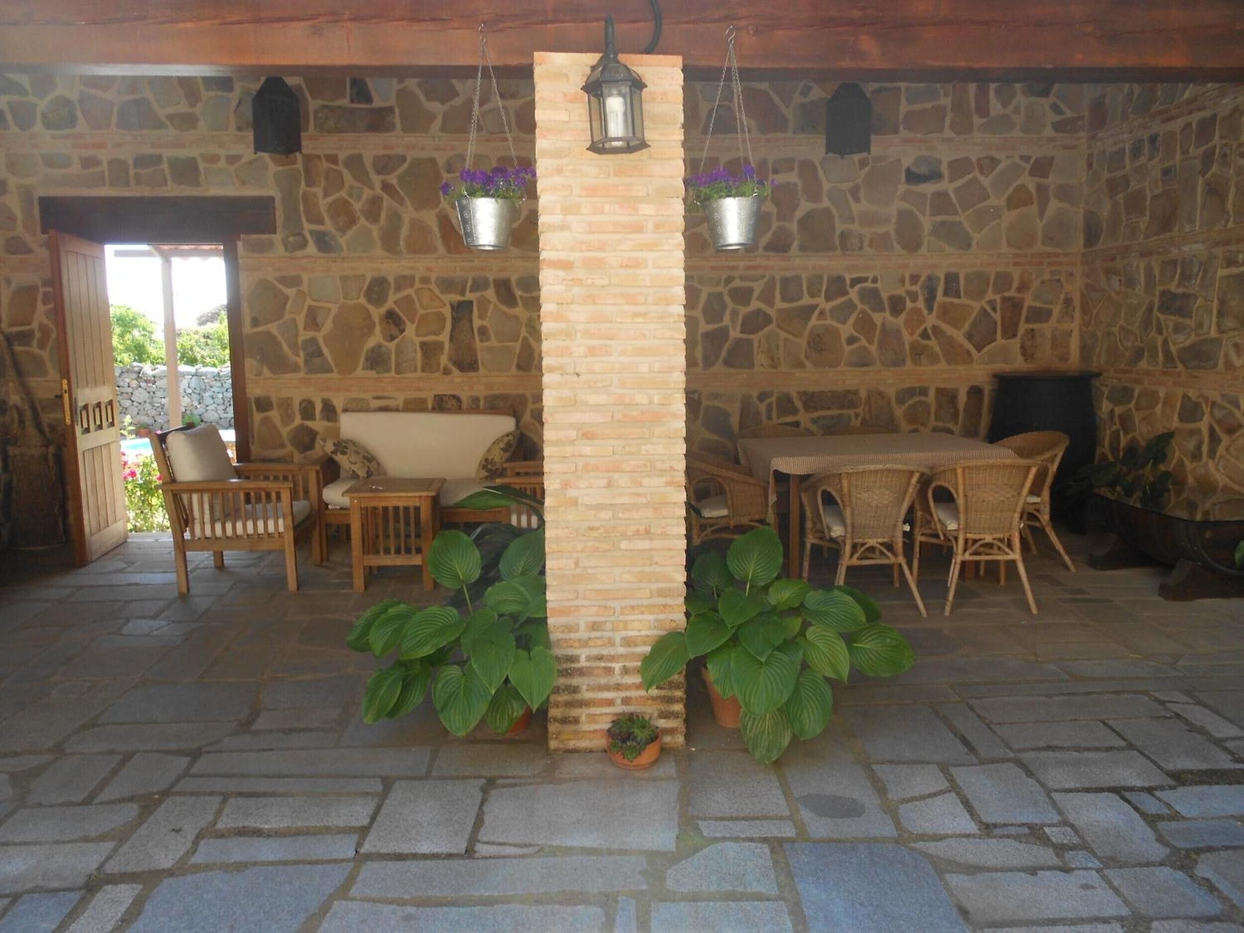 Une terrasse aux murs de pierre, agrémentée de mobilier d’extérieur, d’un pilier central, de lanternes suspendues et de plantes en pot.