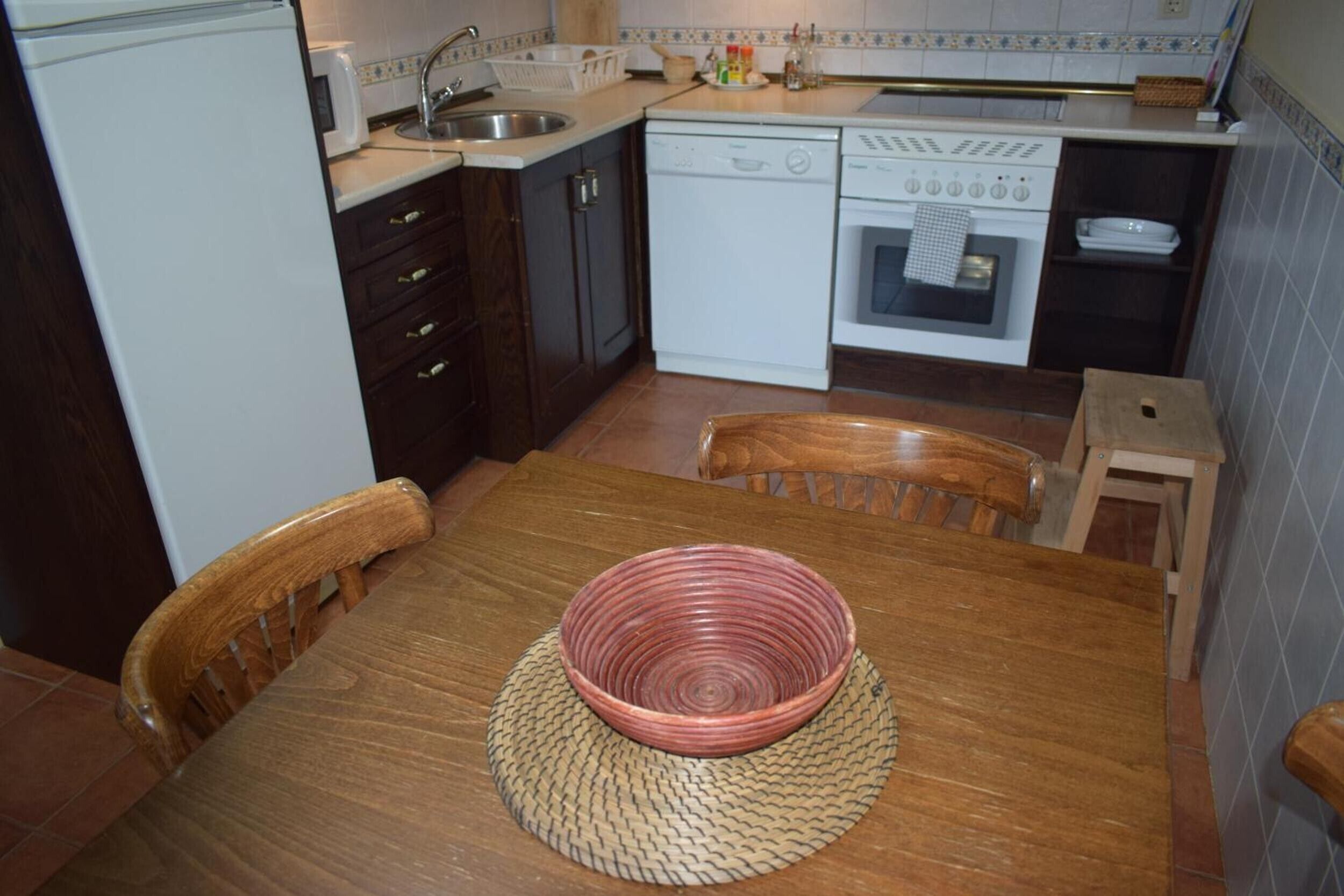 Une cuisine avec une table en bois, des chaises, un saladier rouge, un lave-vaisselle, une cuisinière et un évier.