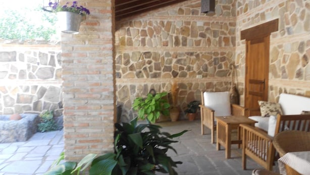 A stone-walled patio with a brick column, potted plants, wicker furniture, and a wooden door. - Casa Rural Las Navillas: vistas a la montaña, Wi-Fi y aire acondicionado (Las Navillas)