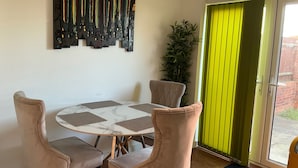 A dining area with a round table, four chairs, a wall-mounted art piece, a potted plant, and a sliding glass door with a green curtain. - House Sleeps 8 With Wifi (Dartford)