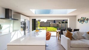 A modern kitchen with a large island, a dining area with a sofa, and a view of a garden. - Stylish Coastal 3BD Bungalow with Garden (West Wittering)