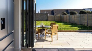 An open sliding door leading to a patio with a table and chairs, a grassy yard, and a wooden fence. - Stylish Coastal 3BD Bungalow with Garden (West Wittering)