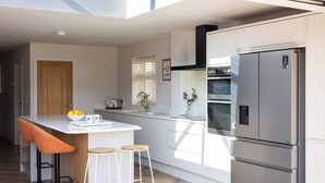 A modern kitchen with a built-in oven, a stainless steel refrigerator, and a bar area with stools. - Stylish Coastal 3BD Bungalow with Garden (West Wittering)