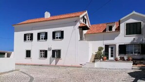 Um edifício branco com telhado de telhas vermelhas, várias janelas com portadas e um pátio pavimentado.