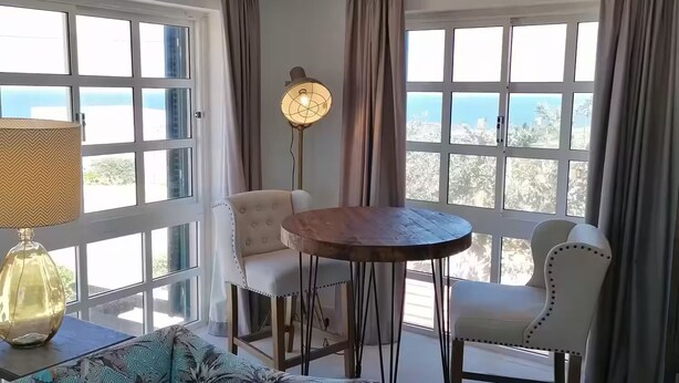 Quarto com sofá, duas cadeiras, mesa, abajur e grandes janelas com cortinas.