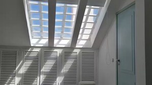 Quarto com claraboias, uma cama, uma porta e um banquinho pequeno.