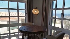 Uma área de refeições com uma mesa redonda, duas cadeiras, um abajur e janelas amplas com cortinas.