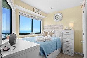 A bedroom with a large window, a bed with a blue blanket, a bedside table with a fan, and a dresser. - T1107 at the Myrtle Beach Resort (Myrtle Beach)