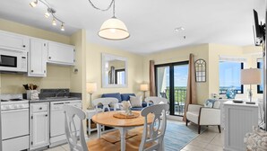 Uma cozinha moderna com armários brancos, uma sala de jantar com mesa de madeira e cadeiras brancas, uma sala de estar com sofá azul e uma varanda com vista.