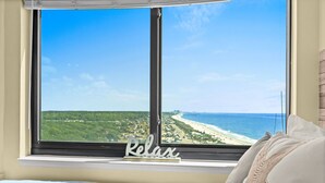 Quarto com uma janela grande dando para uma praia, uma cama com uma toalha azul e uma placa com a palavra 'Relax' no parapeito da janela.