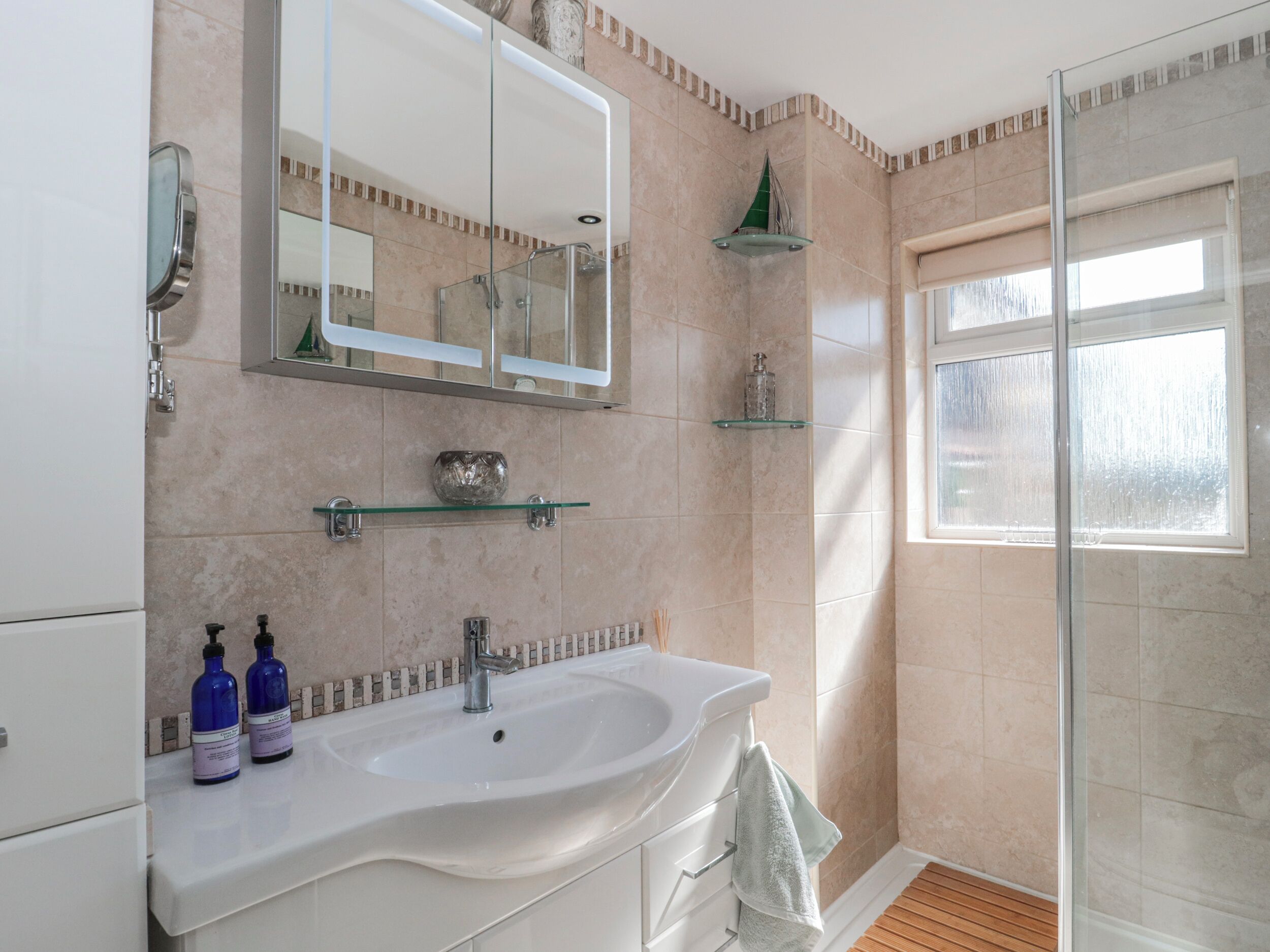 Une salle de bain avec une douche en verre, un grand miroir, un lavabo avec deux bouteilles et une fenêtre.