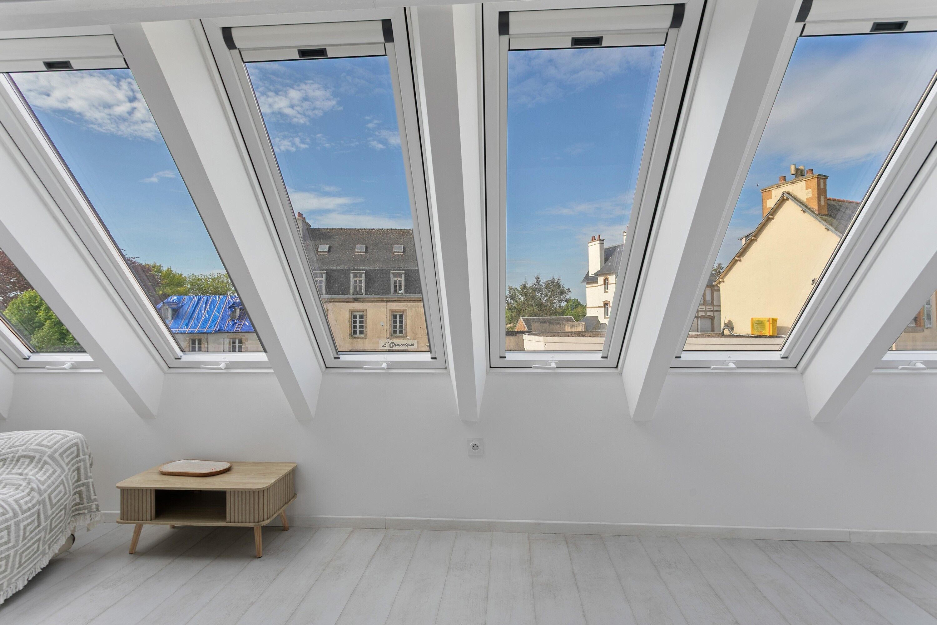 A room with skylights, a bed, a small wooden table, and a view of buildings outside.