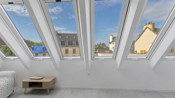 A room with skylights, a bed, a small wooden table, and a view of buildings outside.
