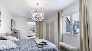 Un dormitorio con cama, un banco, una araña de techo, una ventana con cortinas y un espejo.
