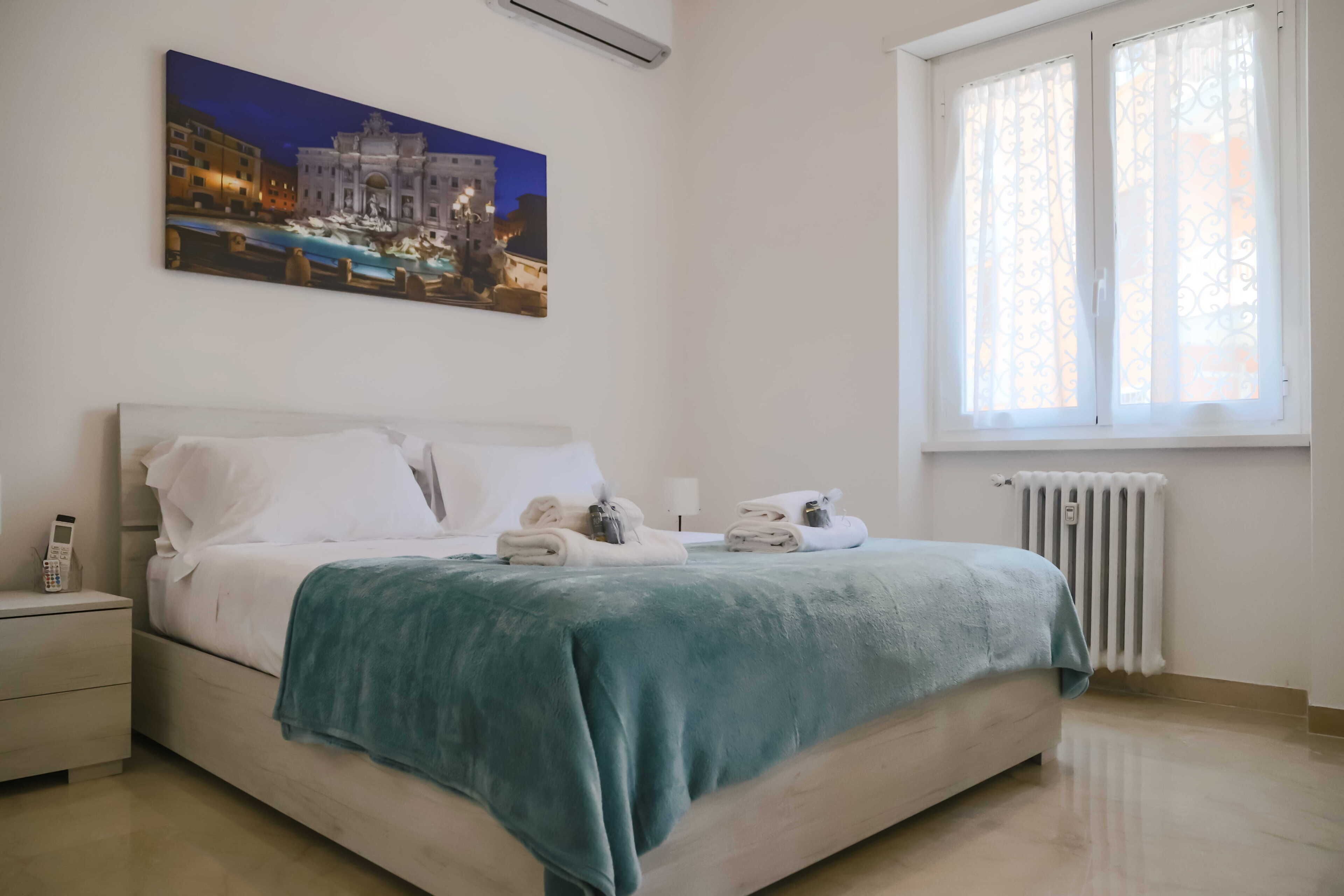 A hotel room with a bed, bedside table, and a painting of a cityscape.