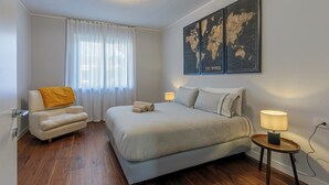 Quarto com cama, uma cadeira, luminária, mapa-múndi na parede e janela com cortinas.
