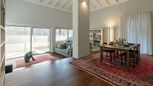 Uma sala de estar ampla com piso de madeira, uma mesa de jantar com cadeiras, uma estante de livros e uma janela grande.