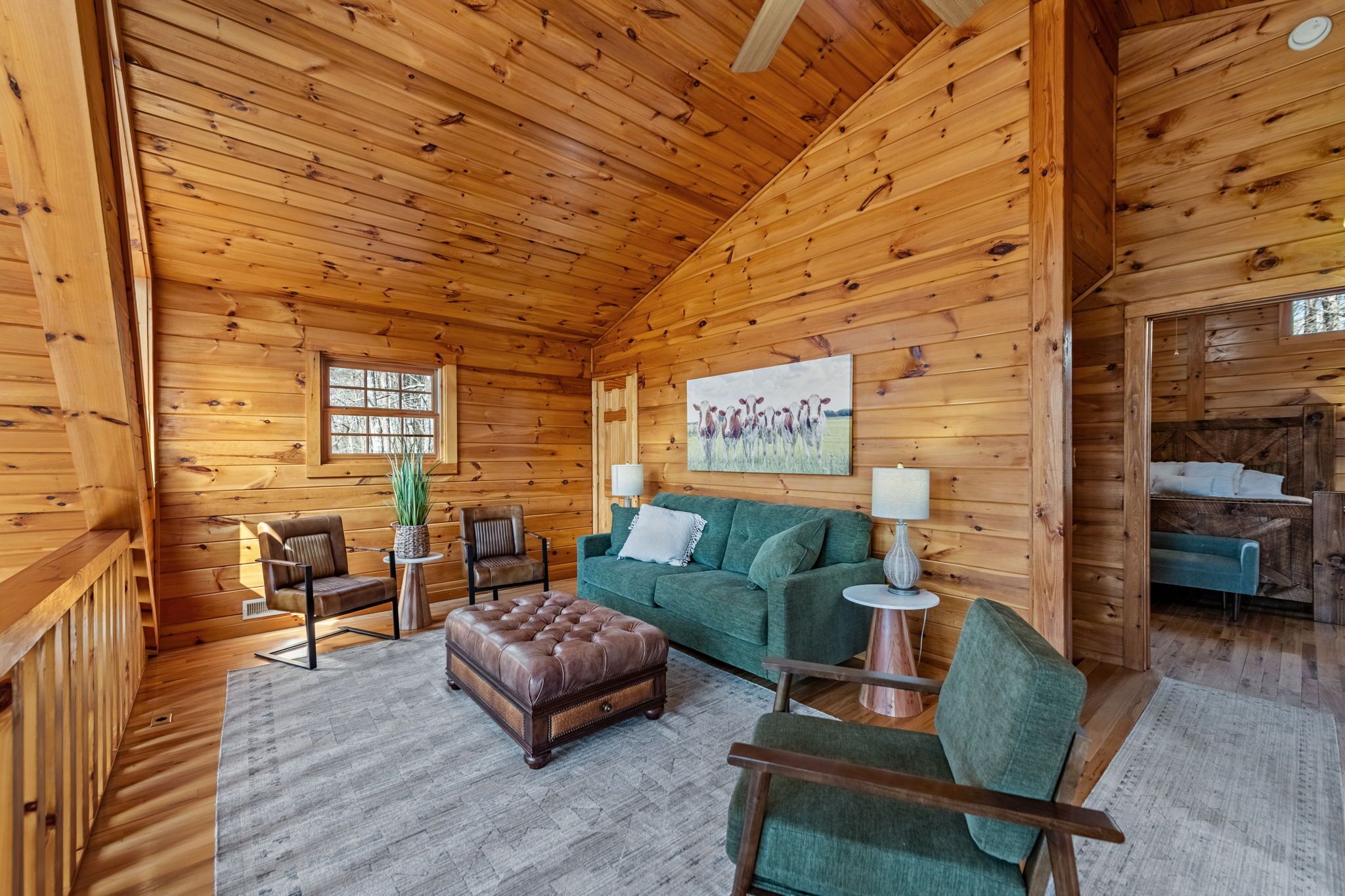 Ein Holzblockhaus-Interieur mit einem grünen Sofa, einem Ledersitzhocker, Holzstühlen, einem Gemälde und einem Fenster.