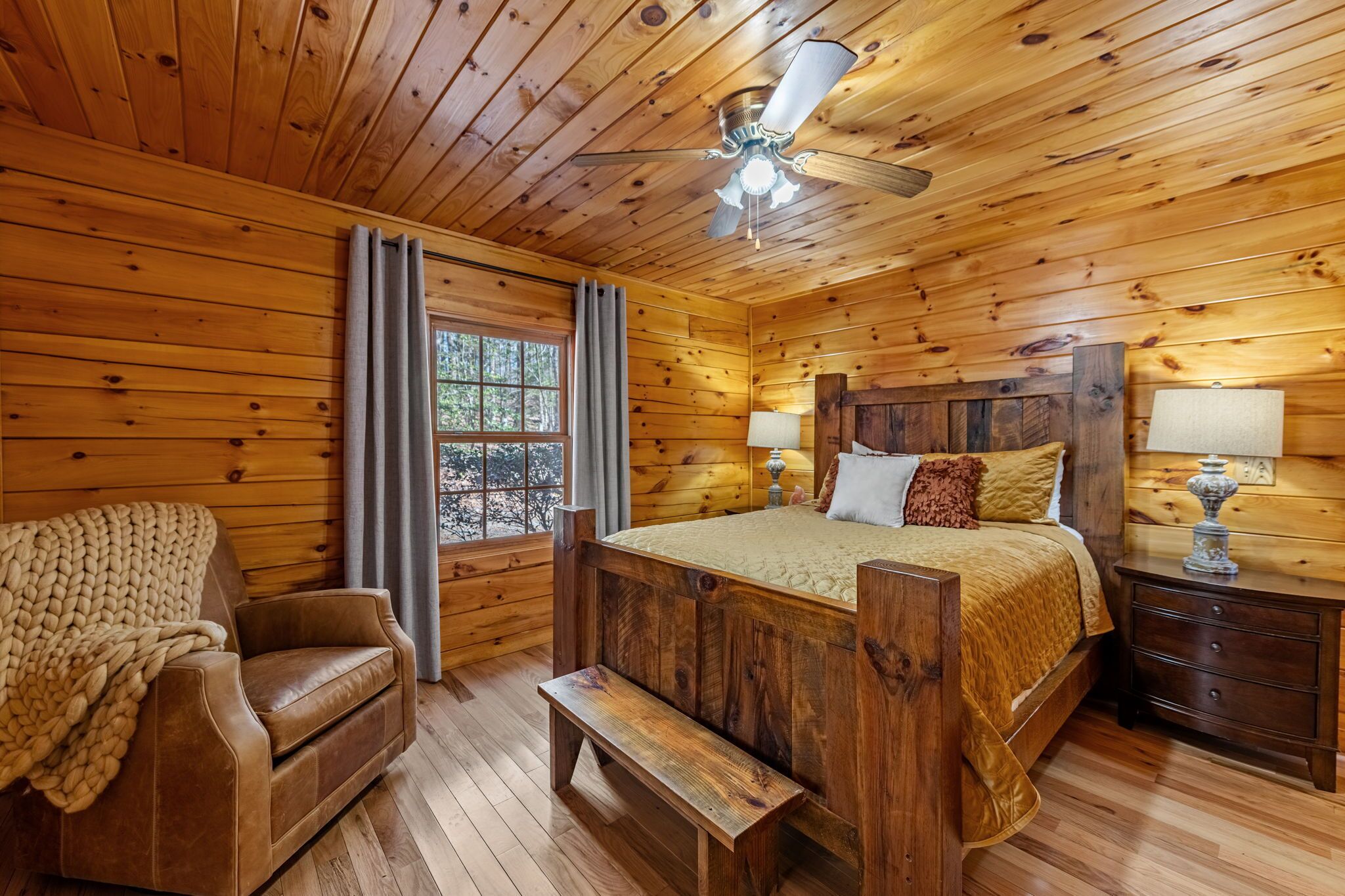 A wooden cabin bedroom with a bed, a chair, a bench, a nightstand, and a ceiling fan.