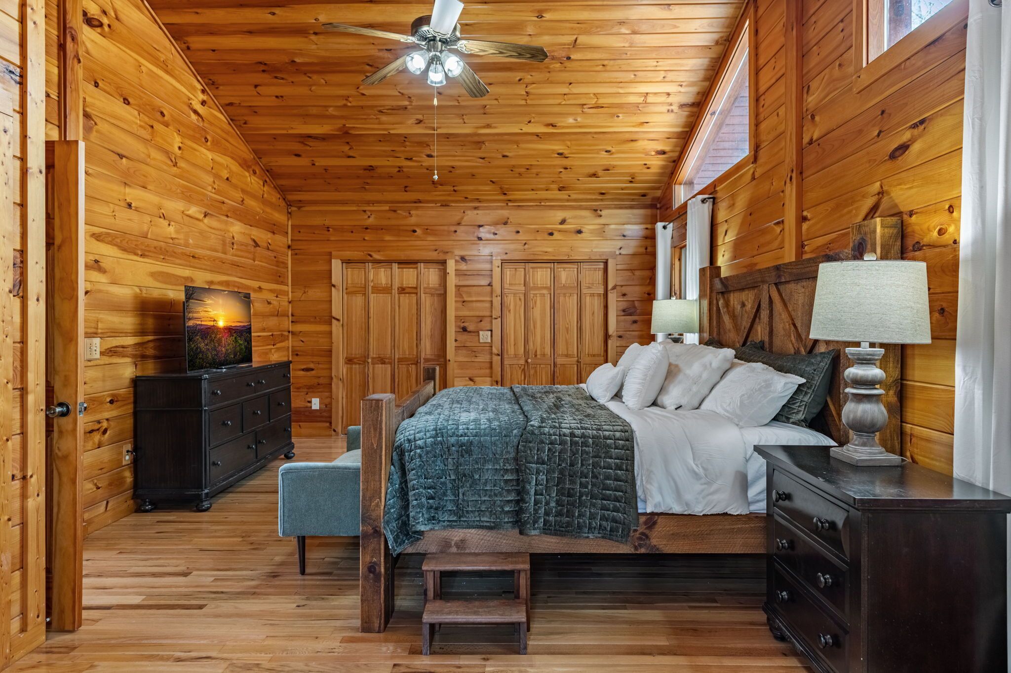 A wooden-paneled bedroom with a bed, dresser, nightstand, and a ceiling fan.