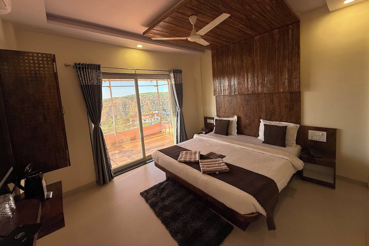 A bedroom with a large bed, a ceiling fan, a view through a sliding door, and a wooden ceiling.