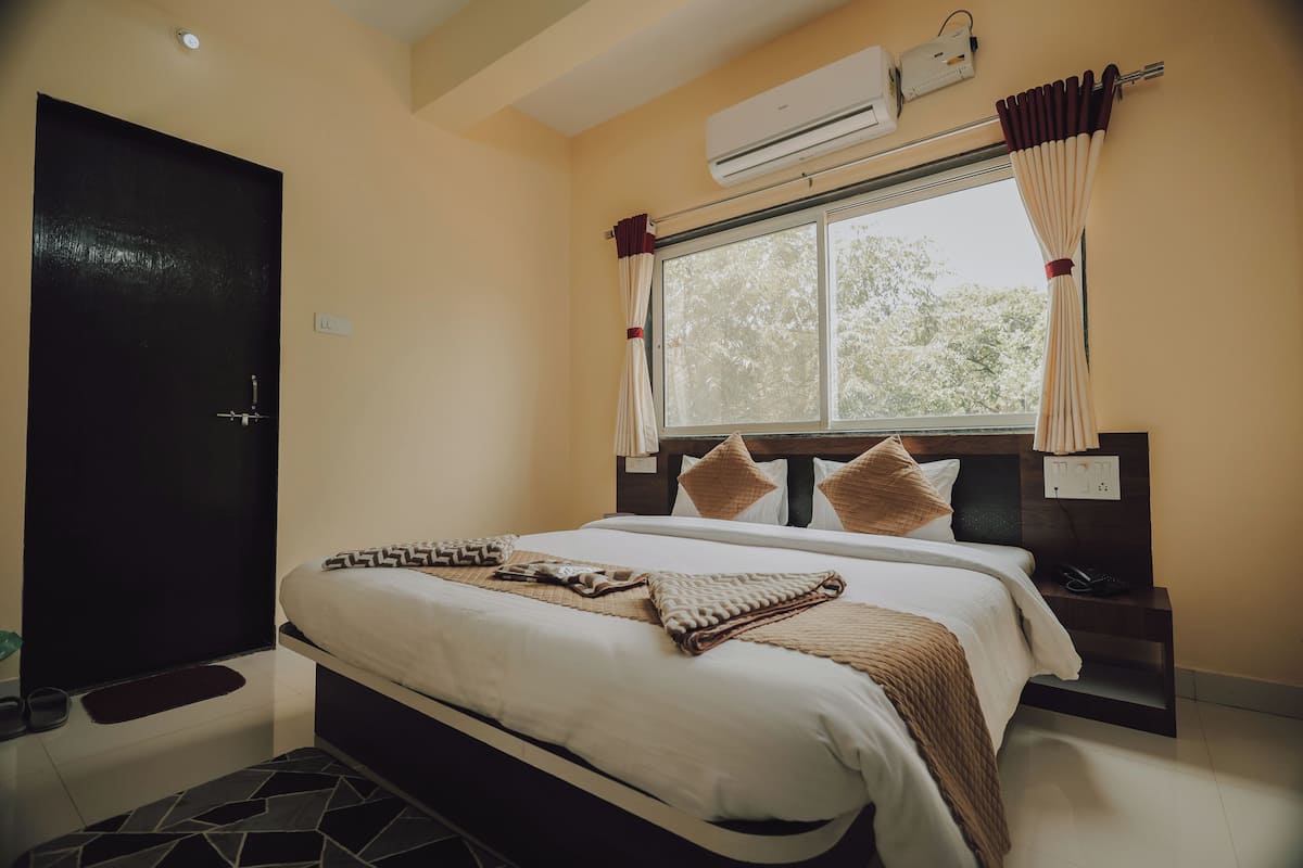 A hotel room with a bed, bedside tables, an air conditioner, a window with curtains, and a door.