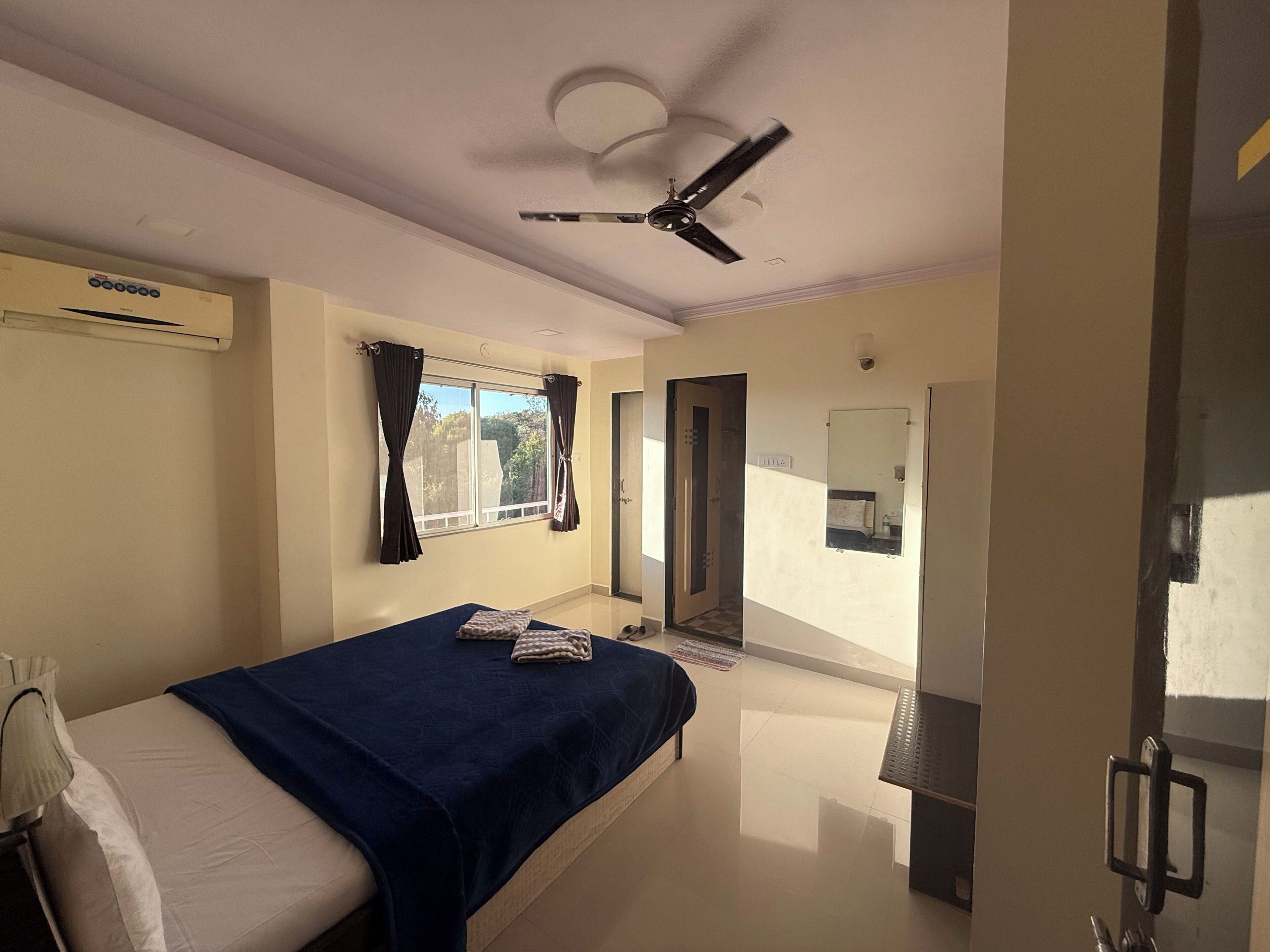 Ein Hotelzimmer mit Bett, Deckenventilator, Fenster mit Vorhängen, Spiegel und einer Tür zu einem anderen Raum.