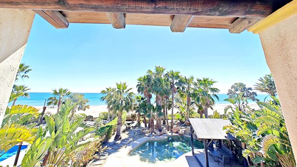 Una pérgola de madera sobre una piscina con vista a una playa y palmeras.