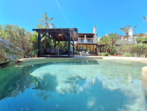 A pool with clear water, a covered seating area with chairs, and a building with balconies in the background.