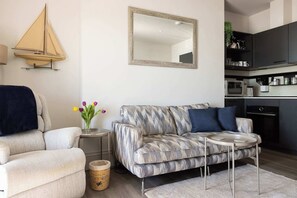 A living room with a sofa, a small table, a mirror, and a kitchen area in the background.